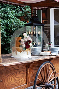 Two tiered white and gold wedding cake with red and pink roses and ruscus leaves on a wooden vintage cart