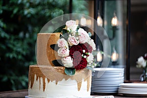 Two tiered white and gold wedding cake with red and pink roses and ruscus leaves on a wooden vintage cart