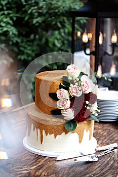 Two tiered white and gold wedding cake with red and pink roses and ruscus leaves on a wooden vintage cart