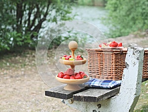 A two-tiered wooden plate with ripe red strawberries, grapes with cheese on skewers and a wicker picnic basket