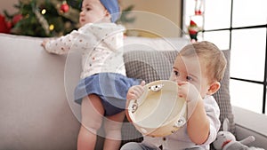 Two toddlers bitting tambourine sitting on sofa by christmas tree at home