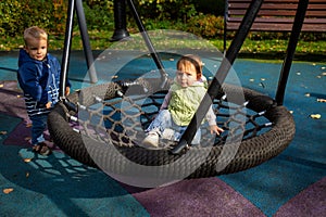 Two toddlers a boy and a girl play cheerfully and swing on a swing hammock on a playground in the autumn park