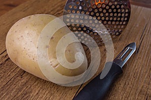A potatoe with a peeler and a tool to crushed potatoe on wooden