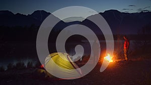 Two tourists camping with tent close to campfire and looking at the mountain.