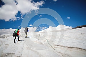 Two tourists, a man and a woman with backpacks and crampons on their feet walk along the glacier against the background