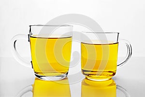 Two transparent glass cups with yellow liquid on white reflective surface. Mugs of tea or juice.