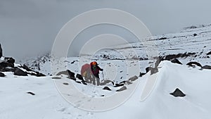 Two travelers with large backpacks are walking along a snow-covered mountain trail.