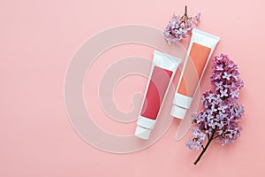 two tubes with cosmetic creams and sprigs of lilac flowers on a pink background