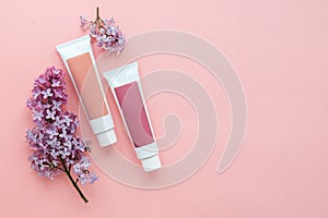 two tubes with cosmetic creams and sprigs of lilac flowers on a pink background