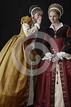 Two Tudor woman whisper together against a black backdrop