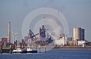 Two tug boats at their moorings with huge steel plant emitting clouds of smoke and wind power stations in the background