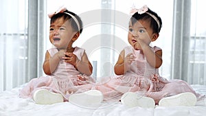 Two twin babies in pink dress on bed, one looking, one crying