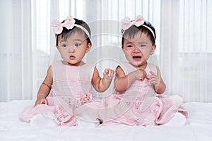 Two twin babies in pink dress on bed, one looking, one crying