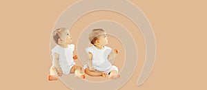 Two twin babies sitting on the floor and looking up together on beige studio background, blank copy space for advertising text