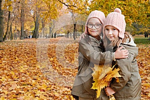Two twin sisters pose for the camera with a bouquet of yellow leaves in the autumn park. Sisters hugging, smiling and