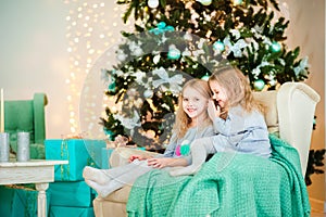 Two twin sisters sitting in a turquoise chair in the hall for the New Year. A sister whispers a secret in her ear in a decorat