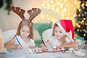 Two twin sisters are writing a letter to Santa Claus for the New Year. A girl with a hat and a girl with reindeer horns