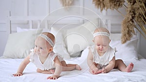 two twins are sitting on a bed in a bright bedroom and smiling, little sister's children are a baby in a cotton