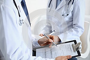 Two unknown doctors filling up medical form on clipboard, just hands closeup. Physicians asking question to patient or