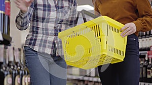 Two unrecognizable caucasian women standing between shelves with alcohol in supermarket and putting bottles of red and