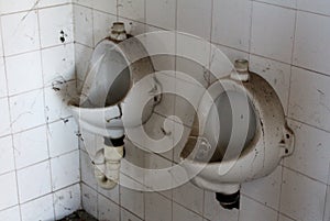 Two vintage retro old disconnected urinals covered with dust and cobweb mounted on white ceramic tiles at abandoned military