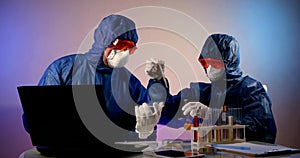 Two virologist doctors in a protective mask and clothing sit at a table with a laptop and test tubes of infection