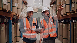 Two warehouse workers wearing hardhats and reflective jackets maintain the detail of manufactured goods in the warehouse