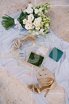 Two wedding rings and bridal accessories on a white fabric with a bouquet of flowers and sandals with heels.