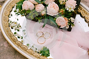 Two wedding rings for the bride and groom and the bride`s engagement ring on a tray with patterns and a bouquet of pink