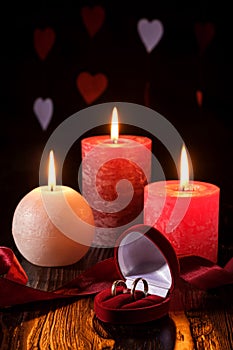 Two wedding rings in red gift box with three wax flame candlelight with ribbon in dark romantic light on hearts background