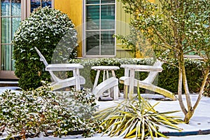 Two white Adirondack chairs with a table between them in the snow in front of a colorful house with green shutters and shrubbery