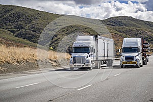 Two white big rigs semi trucks with different loaded semi trailers climbing uphill on the mountain road in California