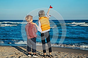 Two white Caucasian children kids, older sister and younger brother playing paper planes on ocean sea beach on sunset