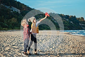 two white Caucasian children kids, older sister and younger brother playing paper planes on ocean sea beach on sunset outdoors