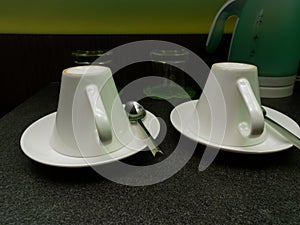 Two white coffee cups and tea spoons put upside down with water glasses and kettle in the background