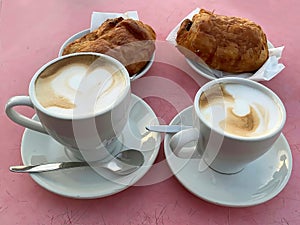 Two white cups of coffee with chocolate croissants on lilac background, selective focus.