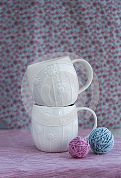 Two white cups stand on top of each other on pink background, Nearby are two tangles for knitting pink and blue