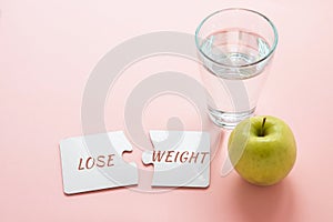 two white disconnected puzzles with the inscription lose weight, a glass of water and a green apple on a pink background