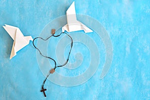 Two white dove origami carrying rosary or scapular in sky blue background.