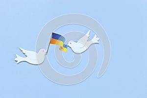 Two white figurines of a doves of peace with  flag of Ukraine  in their beaks on the light blue background. Peace Concept