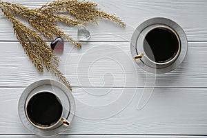 Two white and golden cups of coffee with decorative golden branches and small glass hearts on white wooden background