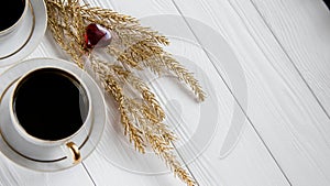 Two white and golden cups of coffee with decorative golden branches and small glass hearts on white wooden background