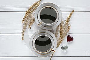 Two white and golden cups of coffee with decorative golden branches and small glass hearts on white wooden background