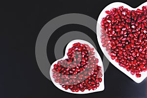 Two white heart shaped plates full of fresh juicy pomegranate seeds, little spoon, whole fruit and ripe one. Toned.