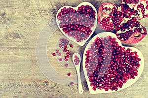 Two white heart shaped plates full of fresh juicy pomegranate seeds, little spoon, whole fruit and ripe one. Toned.