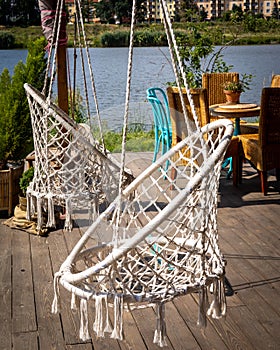 Two white macrame chair swings with tassels on bottom, and a garden furniture on a bright wooden patio, on the riverbank.