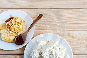 Two white plates with fresh curd and cheesecakes with wooden spoon with plum jam on wooden table with copy space.