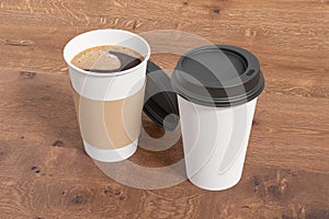 Two white take away coffee paper cups mock up with black lids on wooden background. Opened (with holder) and closed disposable