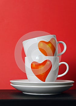 Two white tea cups with painted red hearts on red background, selective focus,copy space