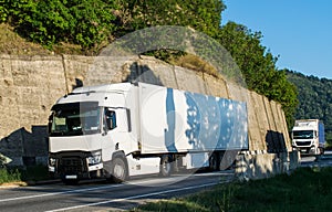 Two white trucks carrying the goods, going on a beautiful road near the mountain